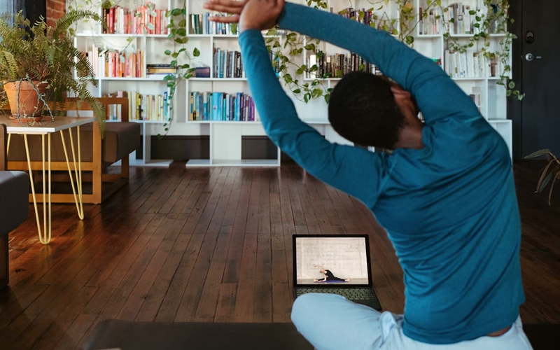 Person seated on mat on floor doing stretching exercises. Wall of books and plants in background.