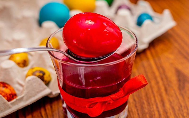 Egg being dyed bright red in foreground; egg carton of different color eggs in background.