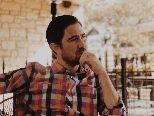 Man in red and black plaid dress shirt seated at table, deep in thought. 