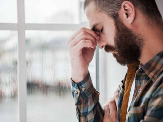 sad young white man with beard standing at window with head bowed and eyes closed