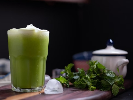 Green liquid in clear drinking glass with an ice cube and fresh greens on the table.