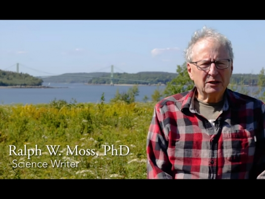 Ralph Moss speaking outside with a green field, water, and bridge in the background