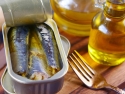 Open tin of sardines on a wood table with a fork and bottle of oil next to it.